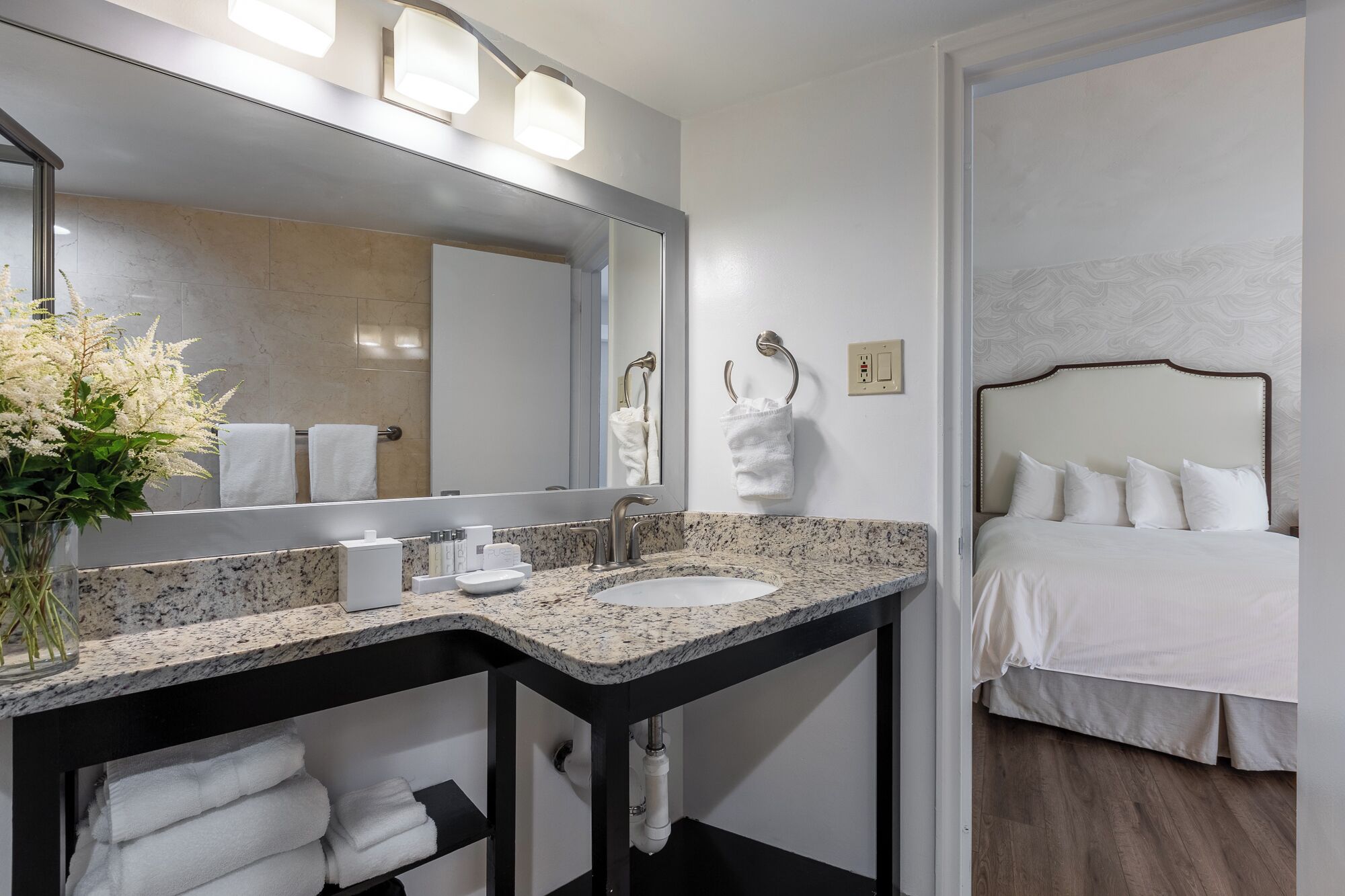 A modern bathroom with a granite countertop, a large mirror, and a bed with white bedding.