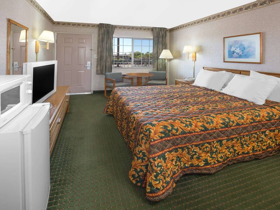 Habitación de hotel con cama, televisor, escritorio con silla, ventana con cortinas y un cuadro en la pared.