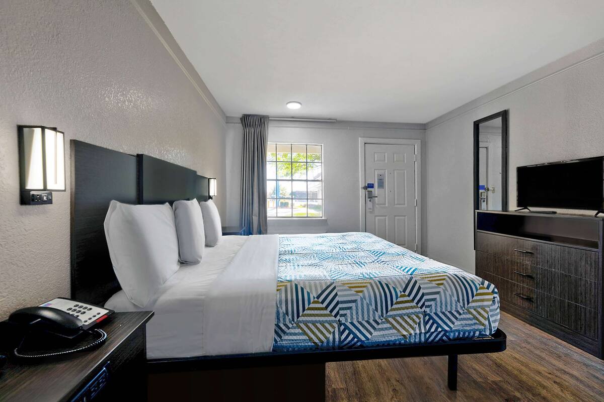 Une chambre d’hôtel avec un lit, une table de chevet avec un téléphone, un téléviseur posé sur un meuble et une fenêtre avec des rideaux.