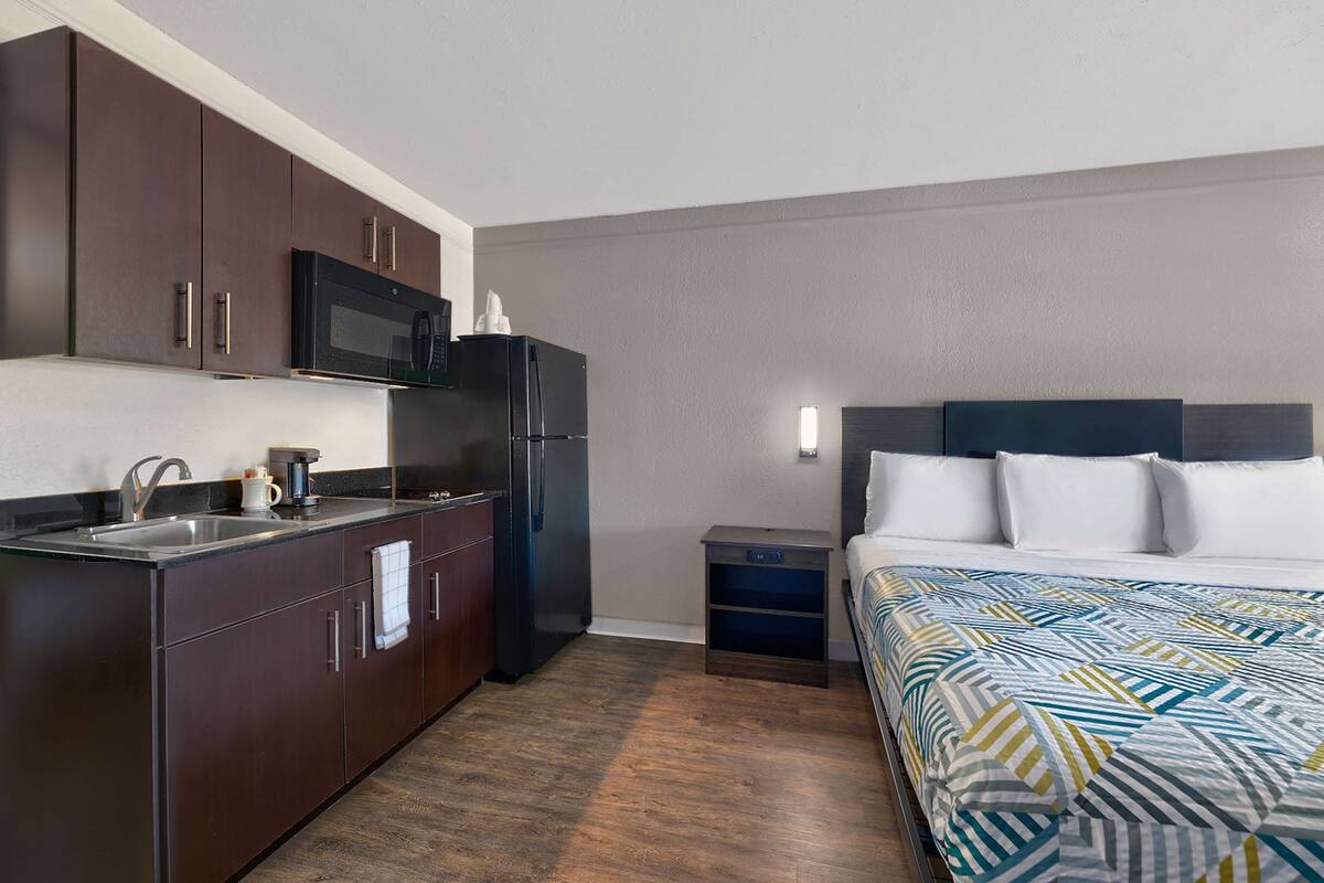 Une chambre d’hôtel compacte dotée d’une kitchenette, comprenant un four à micro-ondes et un réfrigérateur, une petite table de chevet et un lit recouvert d’une courtepointe à motifs.