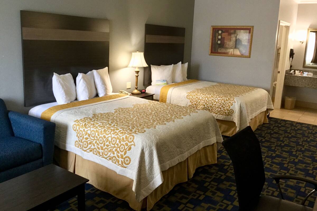 Ein Hotelzimmer mit zwei Betten, einem Schreibtisch, einem Stuhl, einer Lampe, einem Bild und einem durchgegangene Tür erkennbaren Badezimmer.