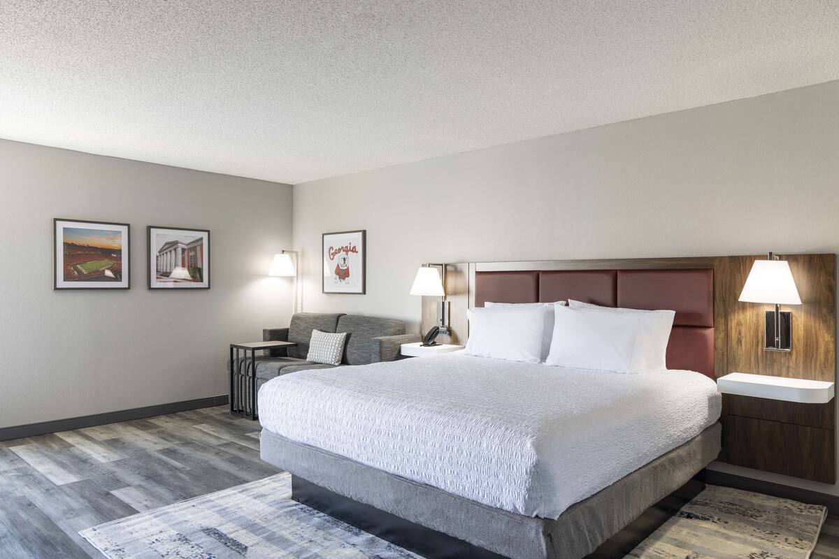 A hotel room with a large bed, a desk, two chairs, a nightstand, and wall-mounted lamps.
