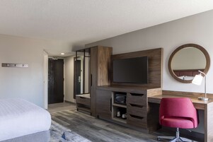A modern hotel room with a bed, a desk with a flat-screen TV, a mirror, and a red chair.
