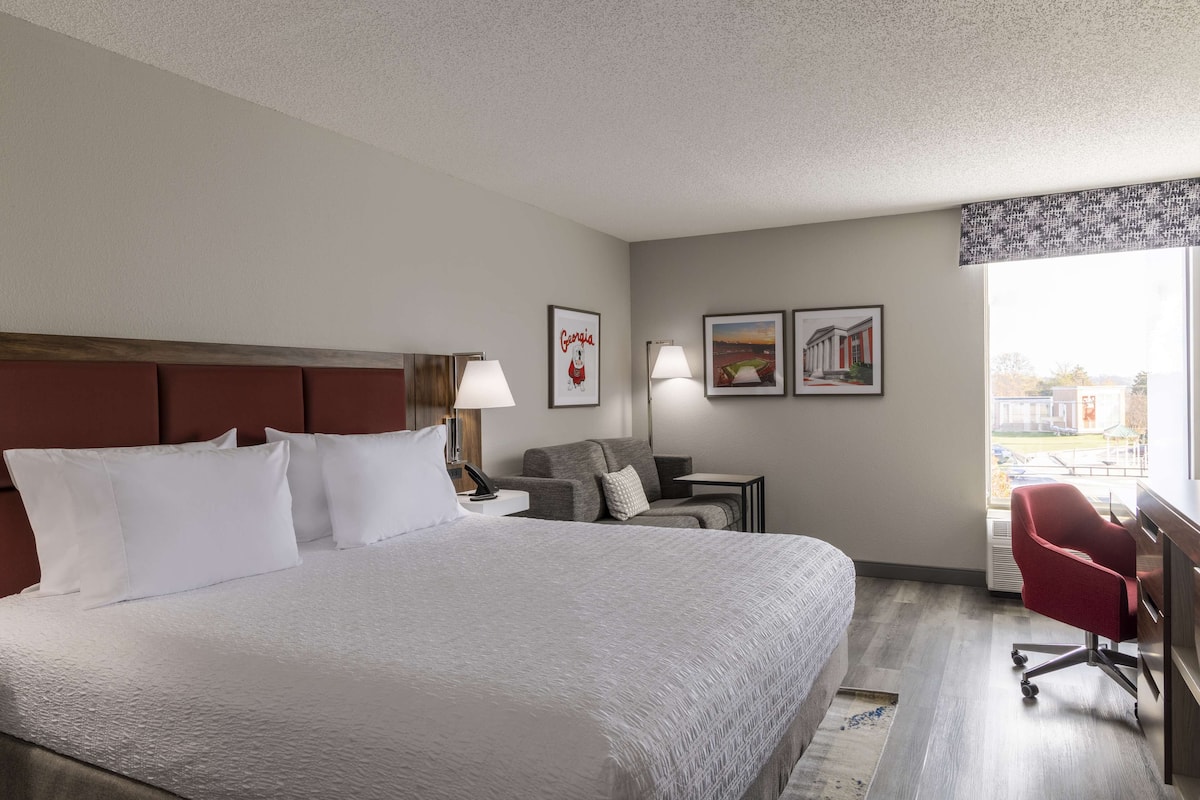 A hotel room with a large bed, a desk, a chair, and a window with a view of buildings.