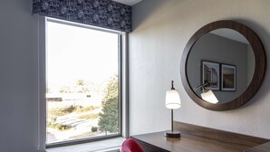 A hotel room with a large window, a round mirror, a desk with a lamp, and a red chair. - Hampton Inn Athens (Athens)