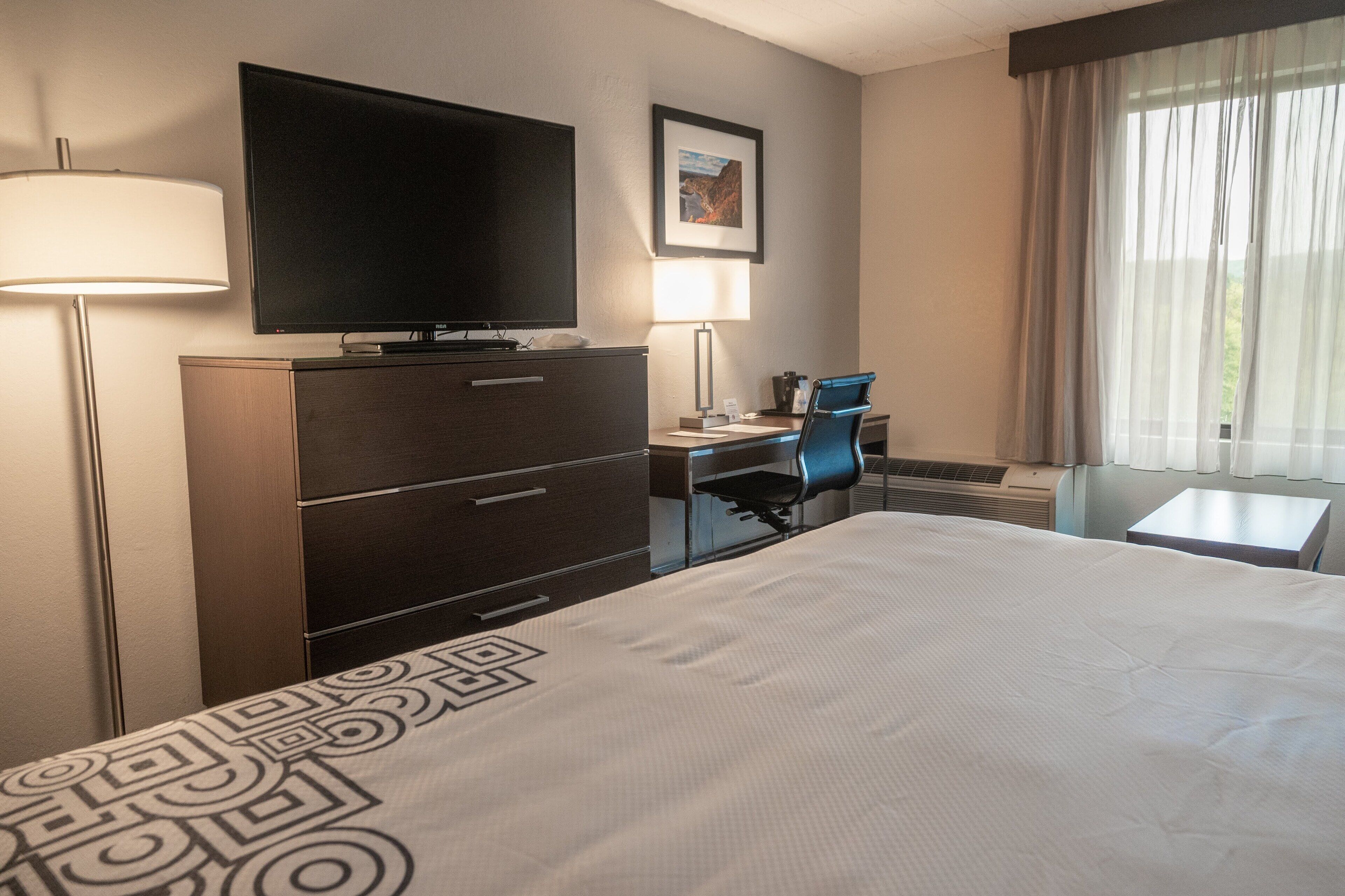 Habitación de hotel con una cama grande, un escritorio con una silla, televisión, lámpara y ventana con cortinas.