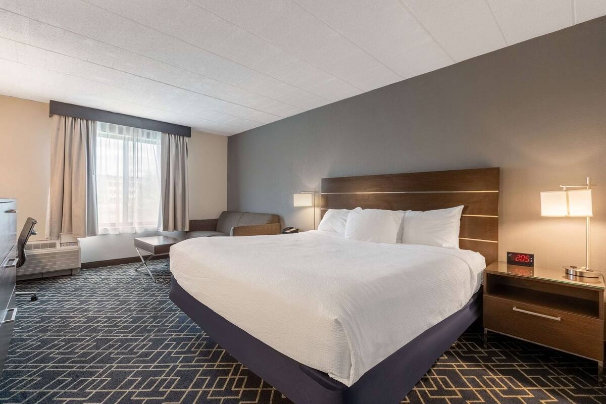 A hotel room with a large bed, a desk, a chair, a nightstand with a lamp, and a window with curtains.