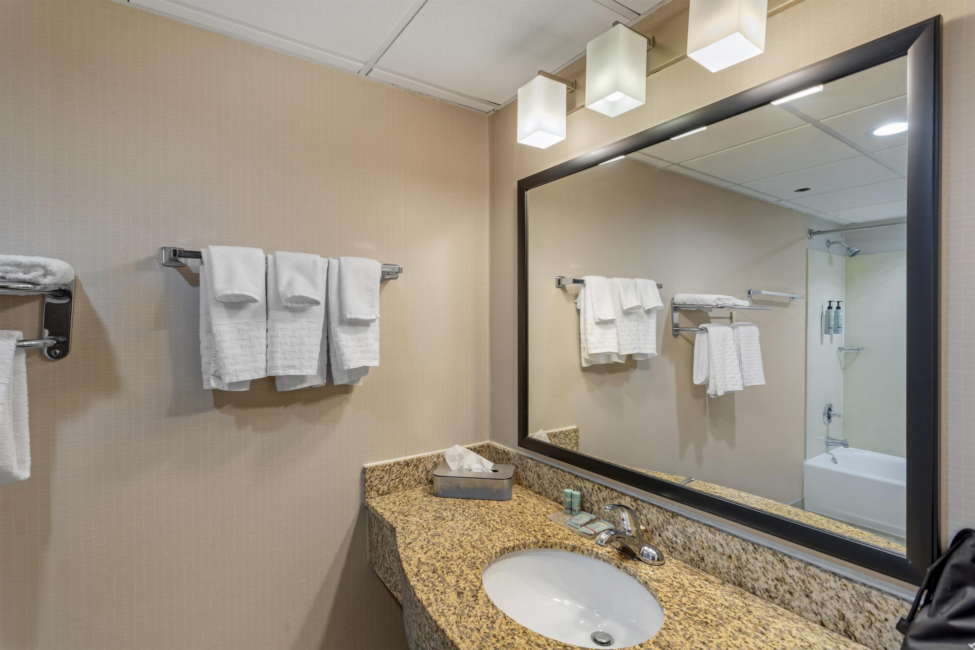 A hotel bathroom with a granite countertop sink, a large mirror, and multiple towel racks.