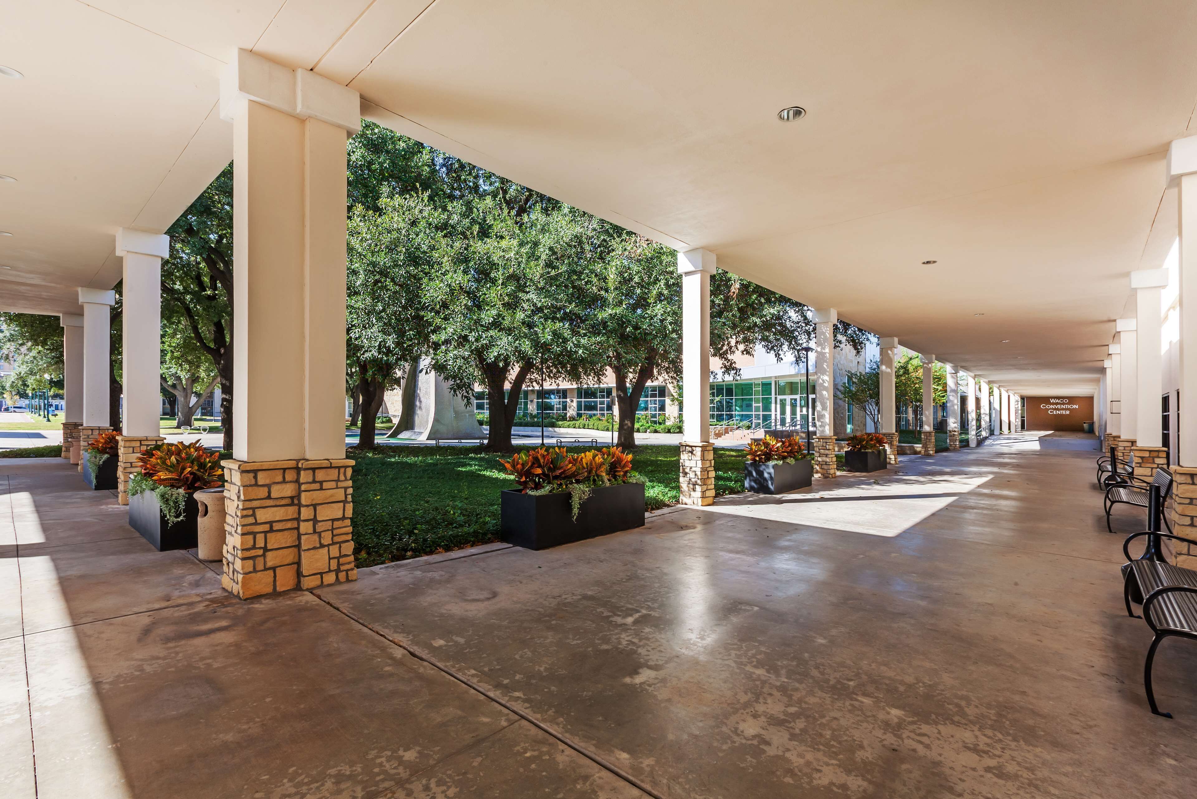 Ett takförsett utomhusområde med pelare, planteringslådor med blommor och en stenlagd gångväg.