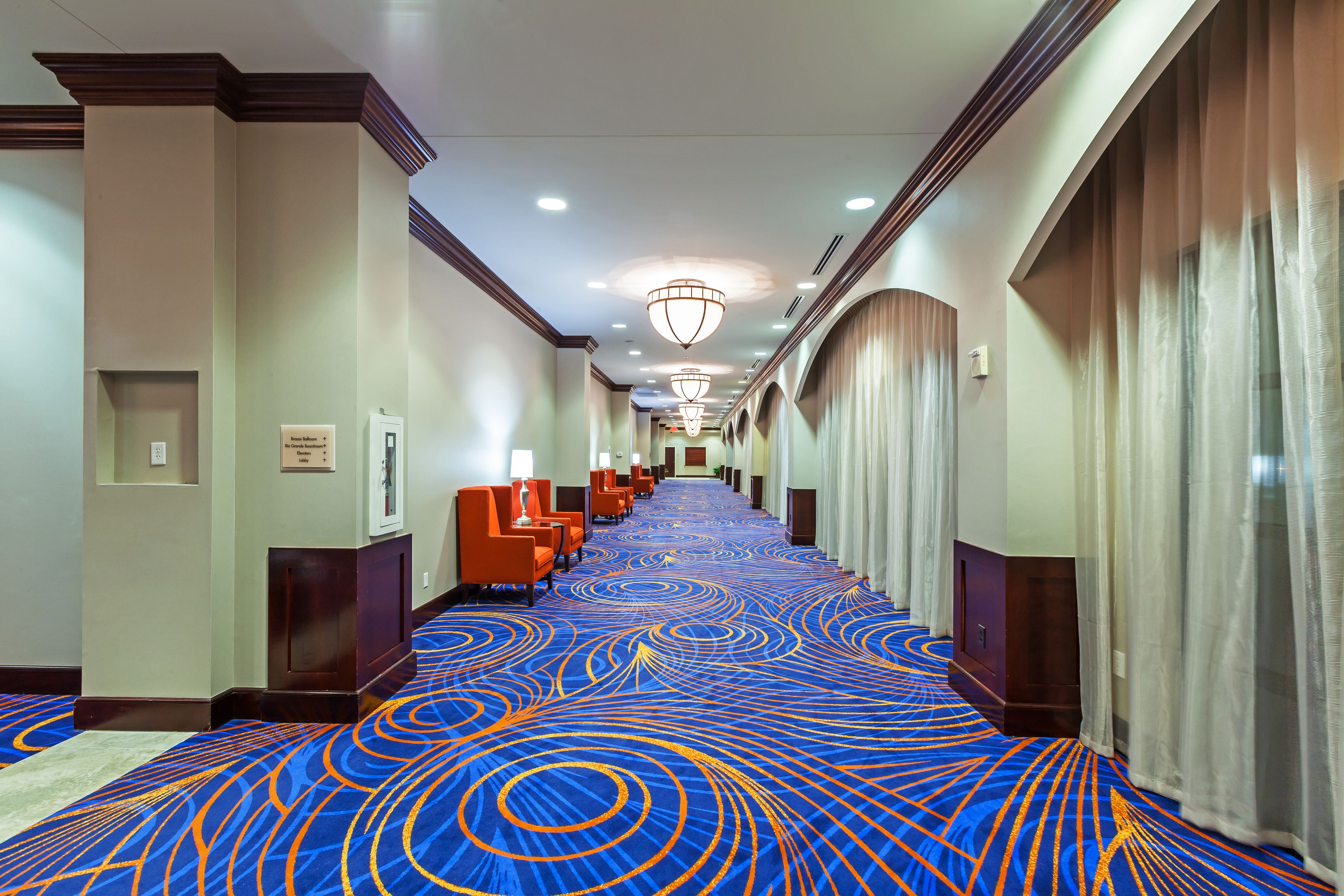 A hotel corridor with a blue carpet, orange chairs, and a chandelier.