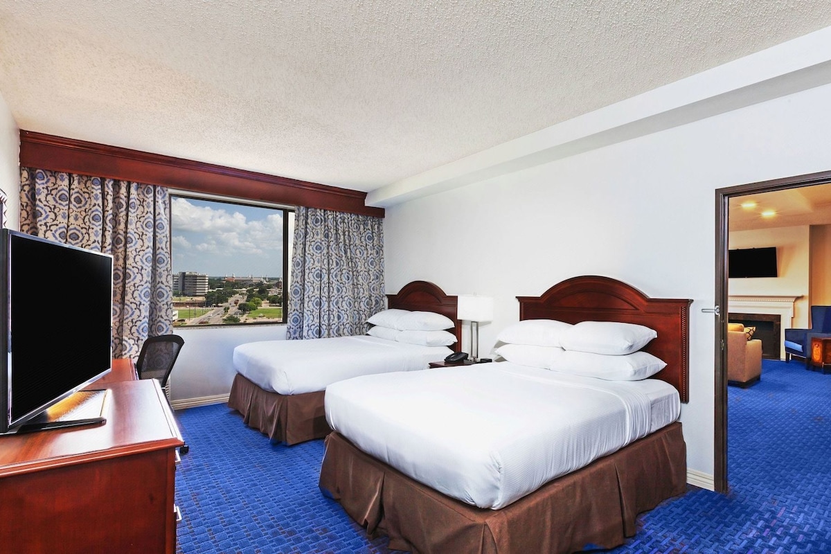 A hotel room with two beds, a desk with a TV, a large window with a view, and a door leading to another room.