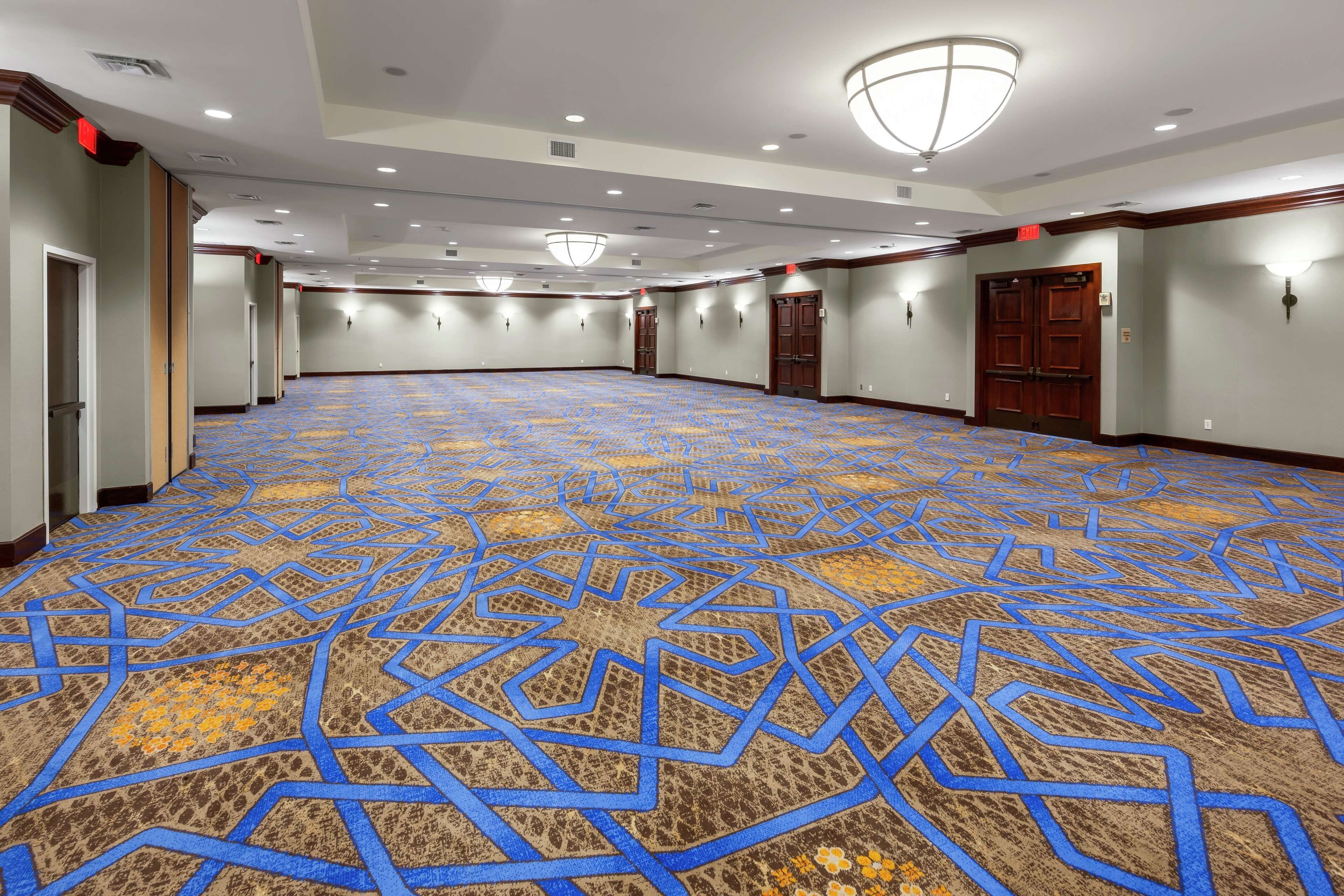 Un amplio salón de conferencias vacío con alfombra estampada, puertas de madera e iluminación empotrada.