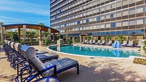 A hotel pool area with lounge chairs, a covered seating area, and a large building with multiple floors. - Hilton Waco (Waco)