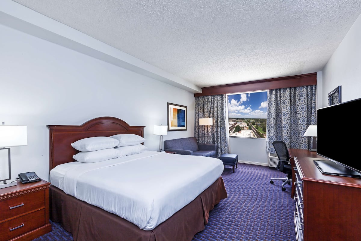 Habitación de hotel con una cama grande, un escritorio con computadora, una silla, un televisor y una ventana con cortinas.