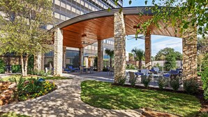 A covered outdoor seating area with stone pillars, surrounded by a landscaped garden and a modern building in the background. - Hilton Waco (Waco)