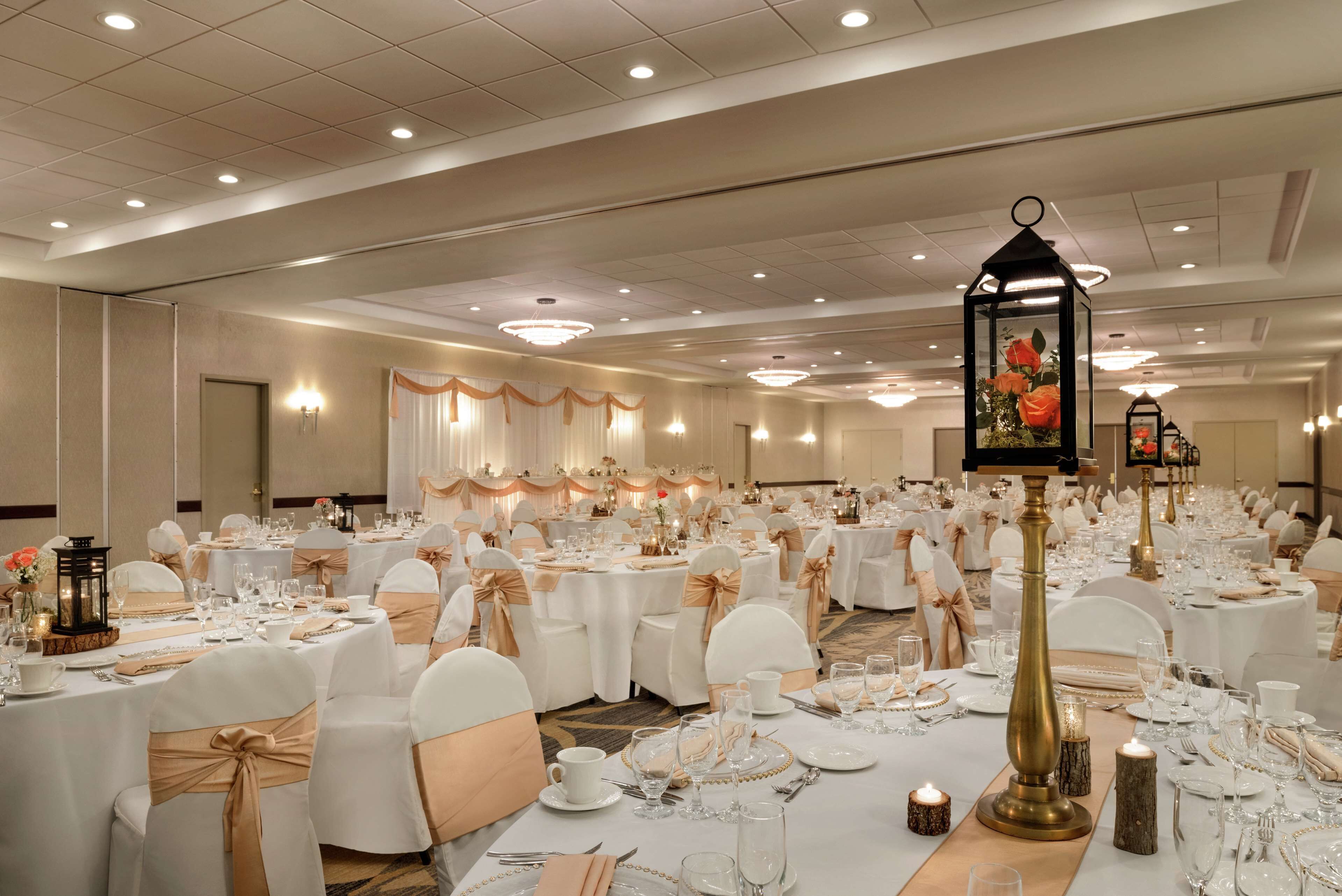 Une salle de banquet avec des tables rondes, des chaises blanches et des rubans dorés. Les tables sont dressées avec des assiettes, des verres et des bougies.