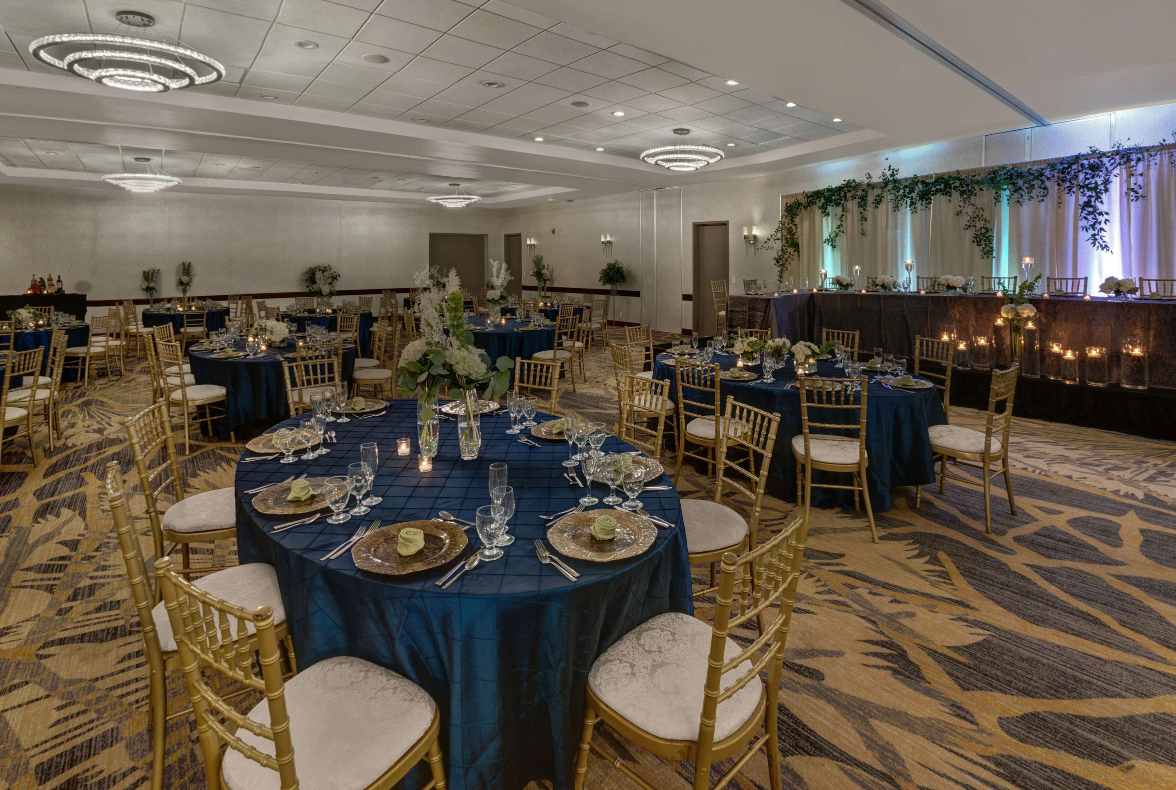 A banquet hall with round tables set for dining, gold chairs, and a decorative wall with greenery.