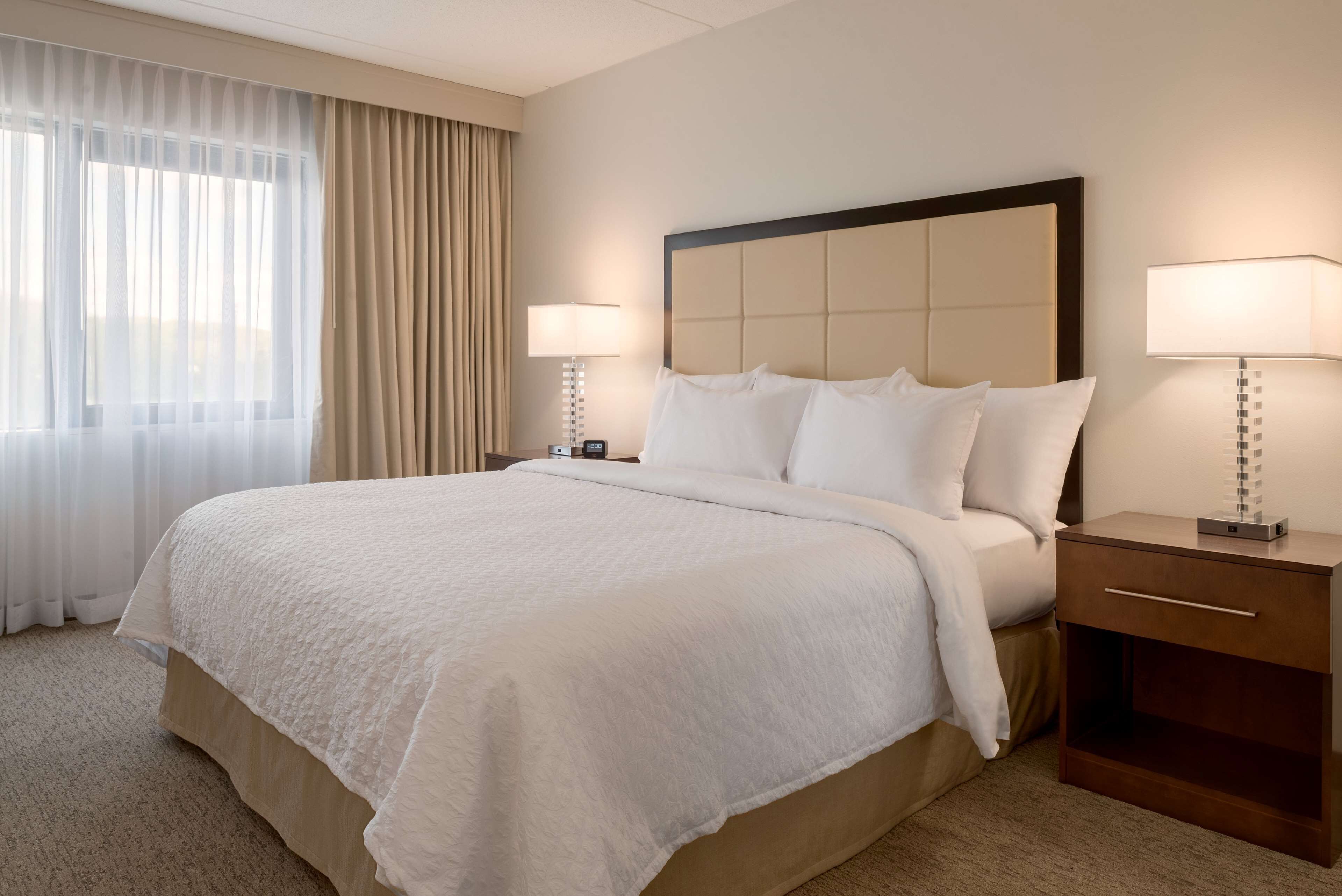 A hotel room with a large bed, two bedside tables with lamps, a nightstand, and a window with sheer curtains.