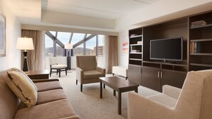 A hotel room with a sofa, armchair, a flat-screen TV, a bookshelf, and a large window offering a city view.