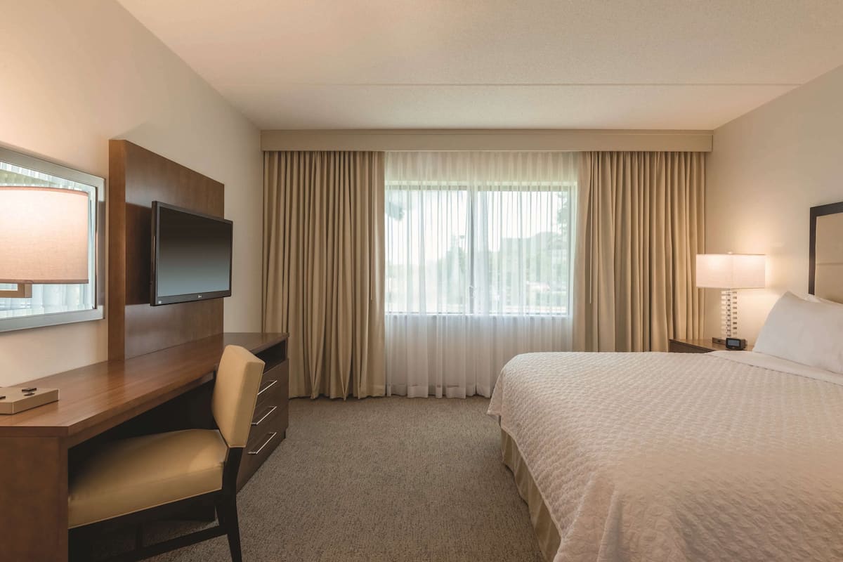 Une chambre d’hôtel avec un lit, un bureau et une chaise, une télévision et une fenêtre avec des rideaux.