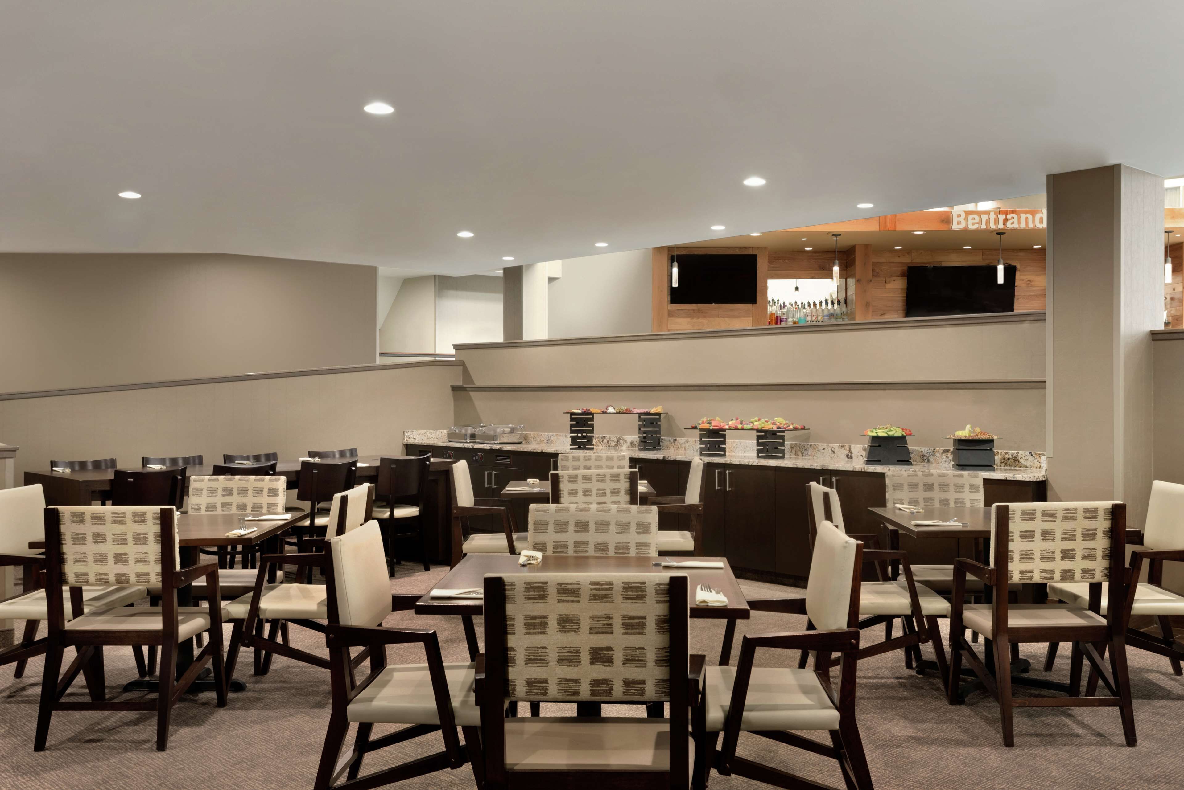 A hotel dining area with multiple tables and chairs, a kitchen area with a counter, and a bar area with a sign.