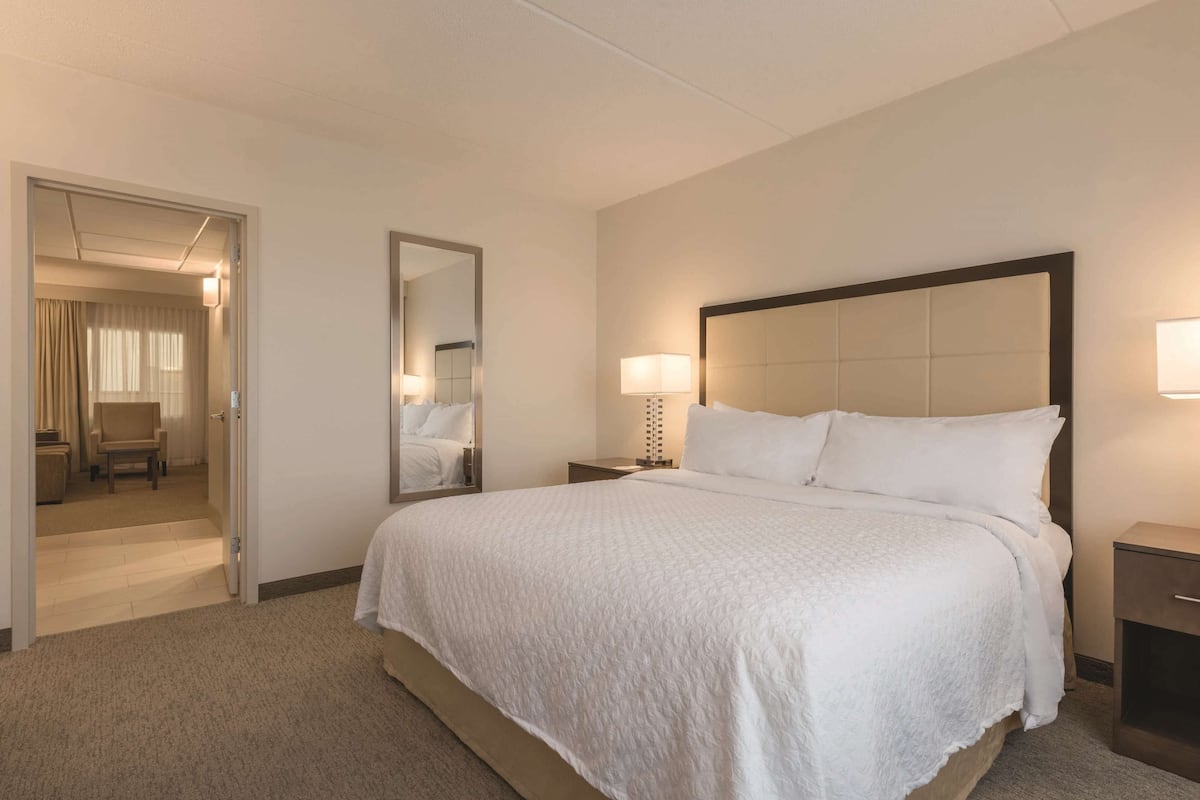 Une chambre d’hôtel avec un grand lit, une table de chevet avec une lampe, un miroir et une porte donnant sur un coin salon avec un fauteuil et des rideaux.