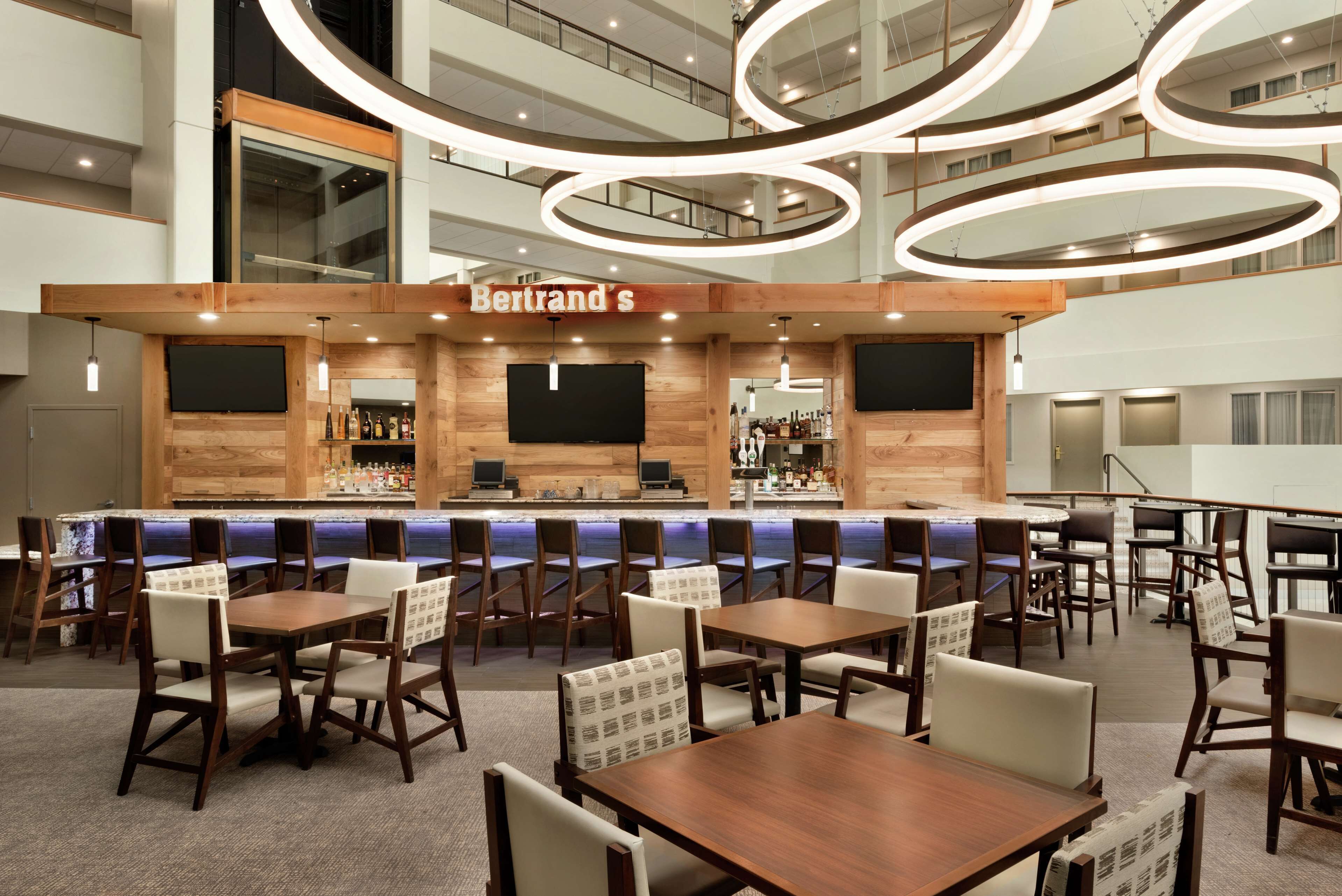 Un moderno bar di hotel con un bancone in legno, diverse aree salotto e un soffitto alto con lampade circolari.