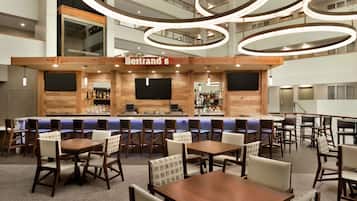 Un moderno bar di hotel con un bancone in legno, diverse aree salotto e un soffitto alto con lampade circolari.