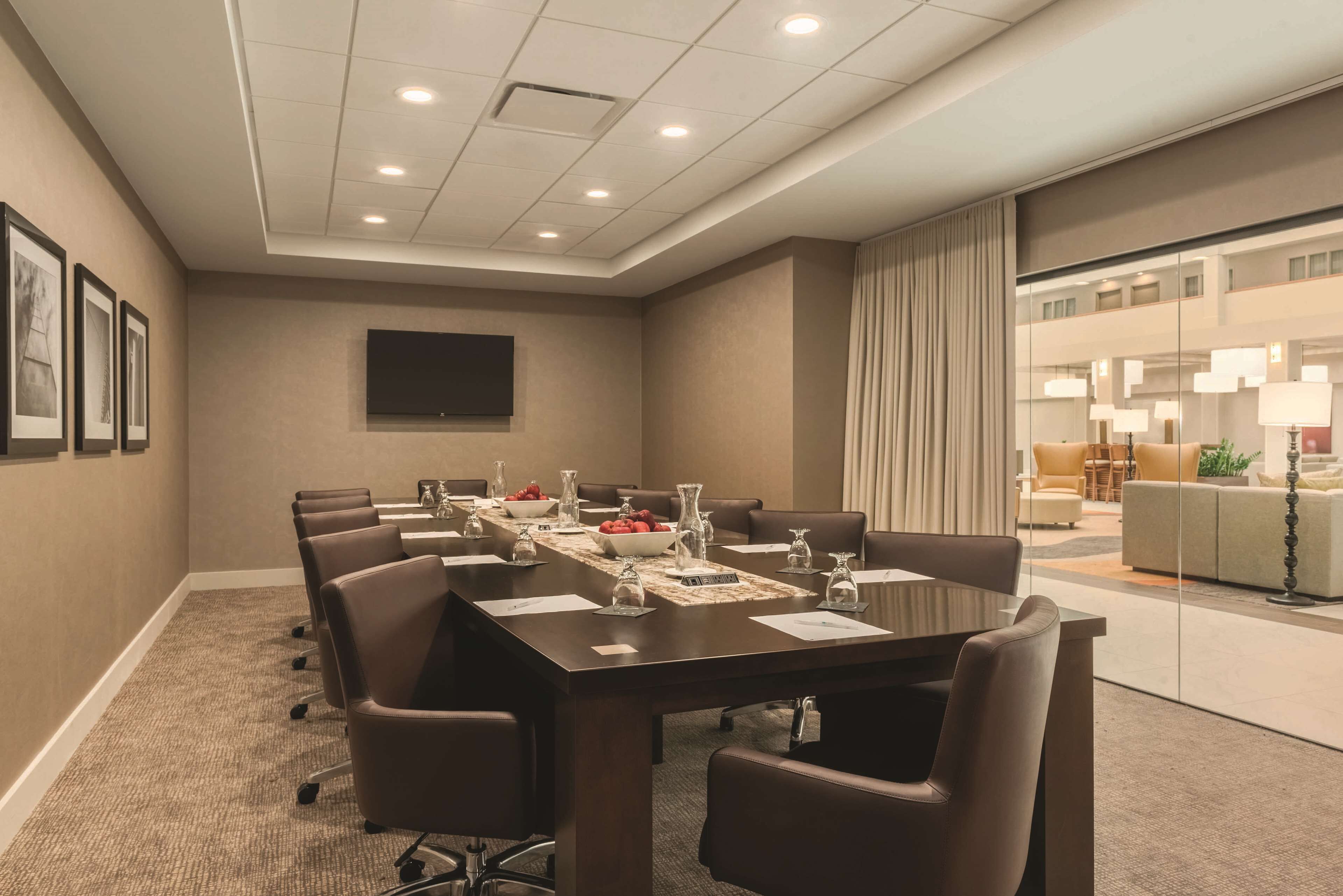 A conference room with a long table, chairs, a flat-screen TV, and a glass partition separating it from a living area with sofas and lamps.