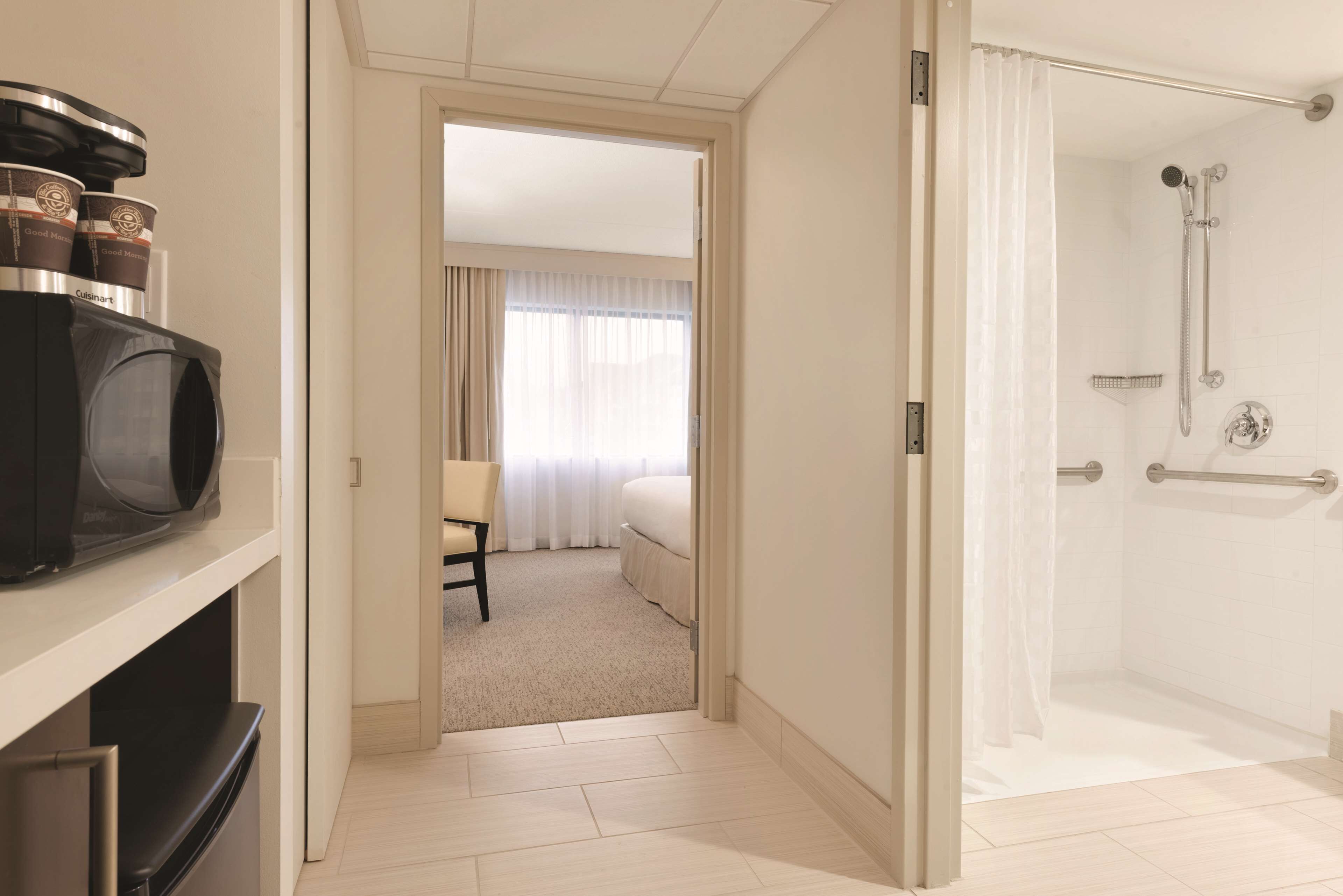 A hotel room with a kitchenette, a shower area, and a view of a bedroom with a bed and curtains.