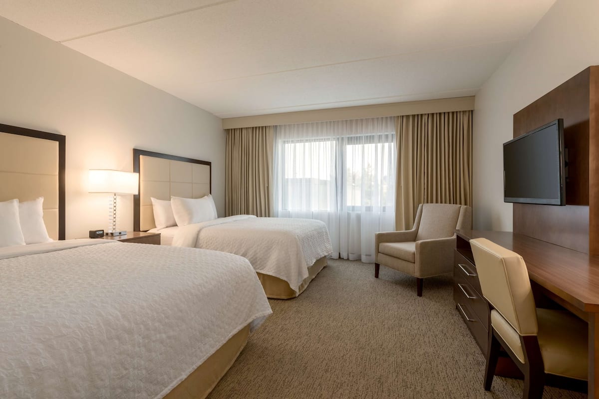 Une chambre d’hôtel avec deux lits, un bureau, une chaise, une télévision et une grande fenêtre avec des rideaux.