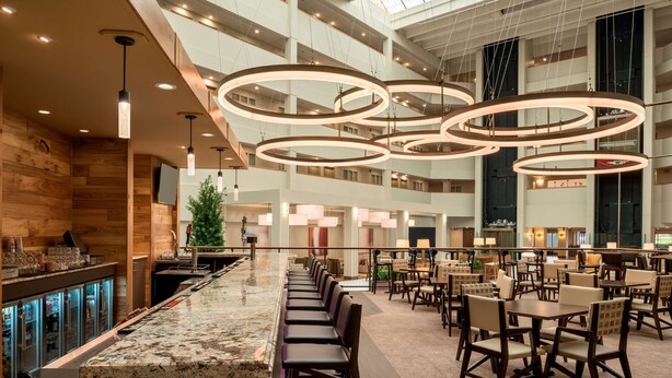 A spacious hotel lobby with a high ceiling, circular lighting fixtures, a central seating area, and a bar with a granite countertop.