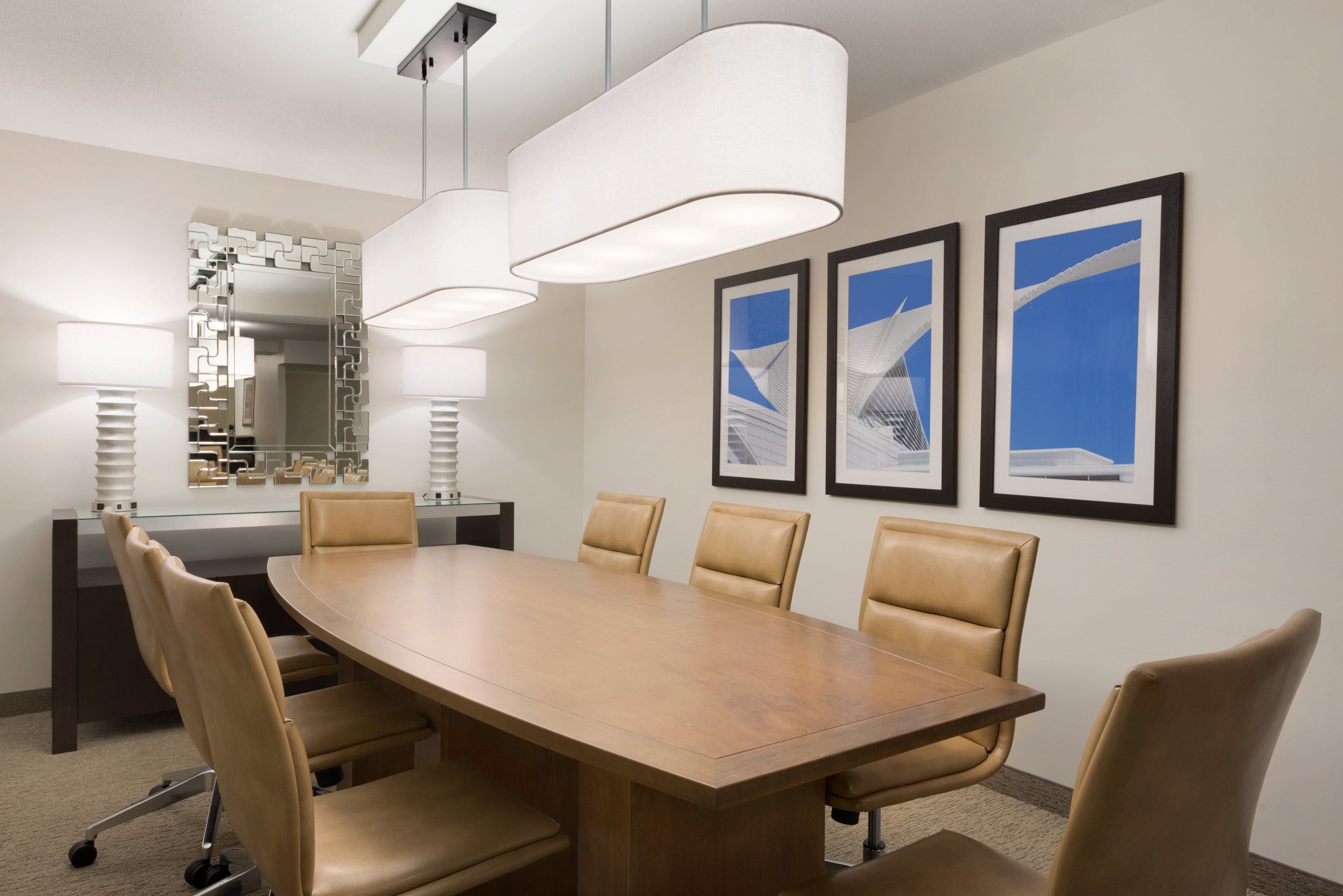 A conference room with a large wooden table, six leather chairs, two pendant lights, a glass partition, a mirror, and three framed pictures on the wall.