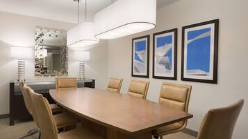 A conference room with a large wooden table, six leather chairs, two pendant lights, a glass partition, a mirror, and three framed pictures on the wall.