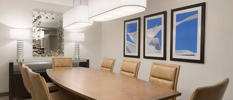 A conference room with a large wooden table, six leather chairs, two pendant lights, a glass partition, a mirror, and three framed pictures on the wall.