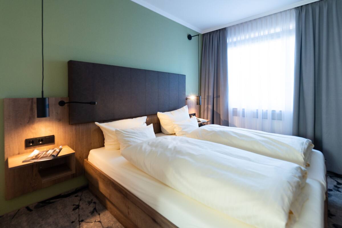 Una cama doble con ropa blanca, un cabecero de madera, una mesita de noche con lámpara y una ventana con cortinas.
