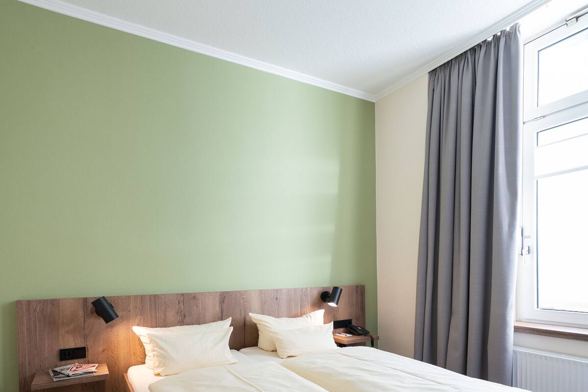 Cama doble con cabecera de madera, dos mesitas de noche con lámparas, una ventana con cortinas y una pared verde.