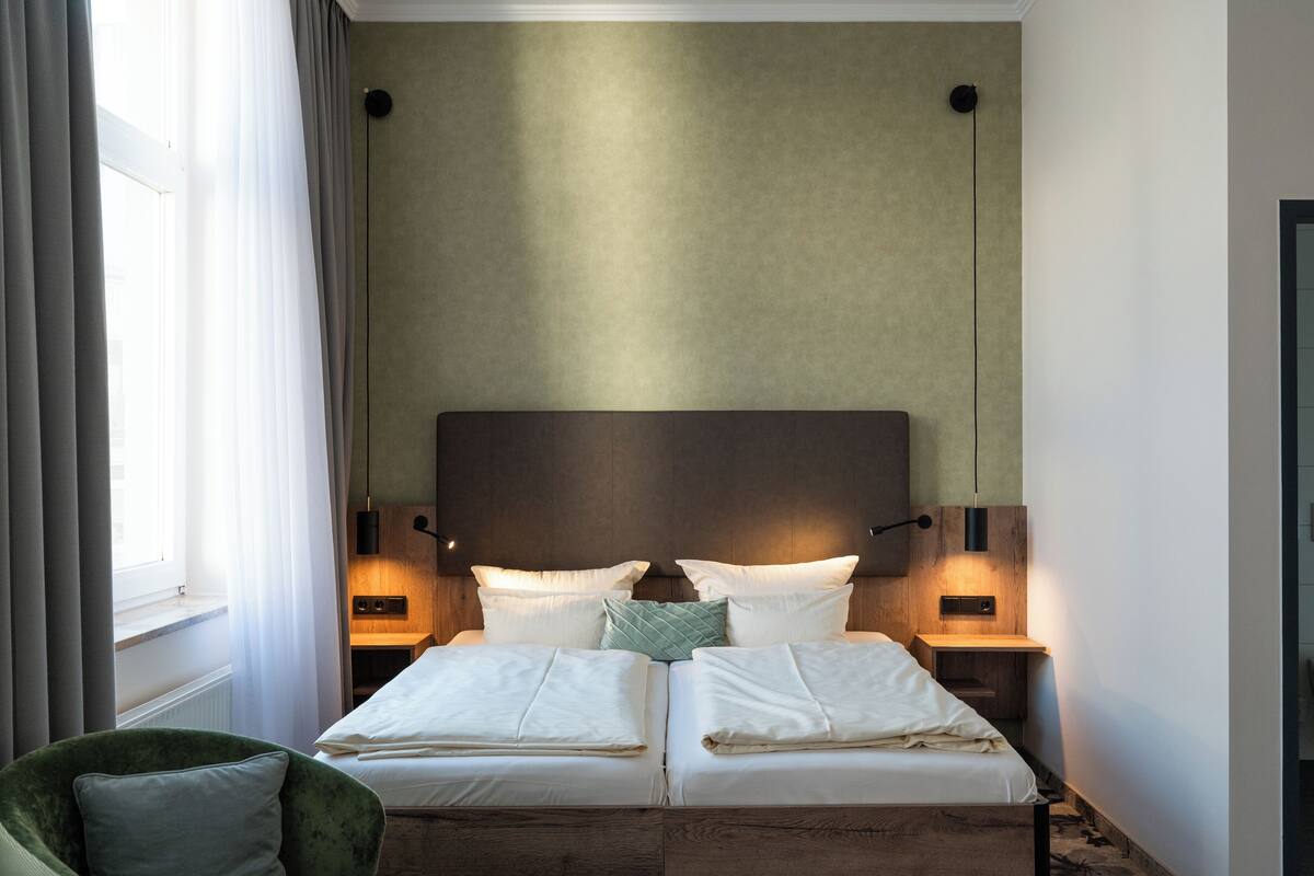 Una cama doble con dos mesitas de noche, una silla verde y un ventanal con cortinas.
