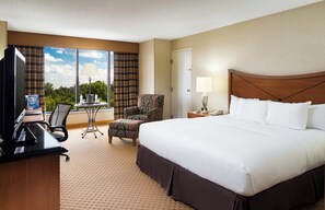 A hotel room with a large bed, a desk with a computer, a chair, a small table with a lamp, and a view of the outdoors through the window.