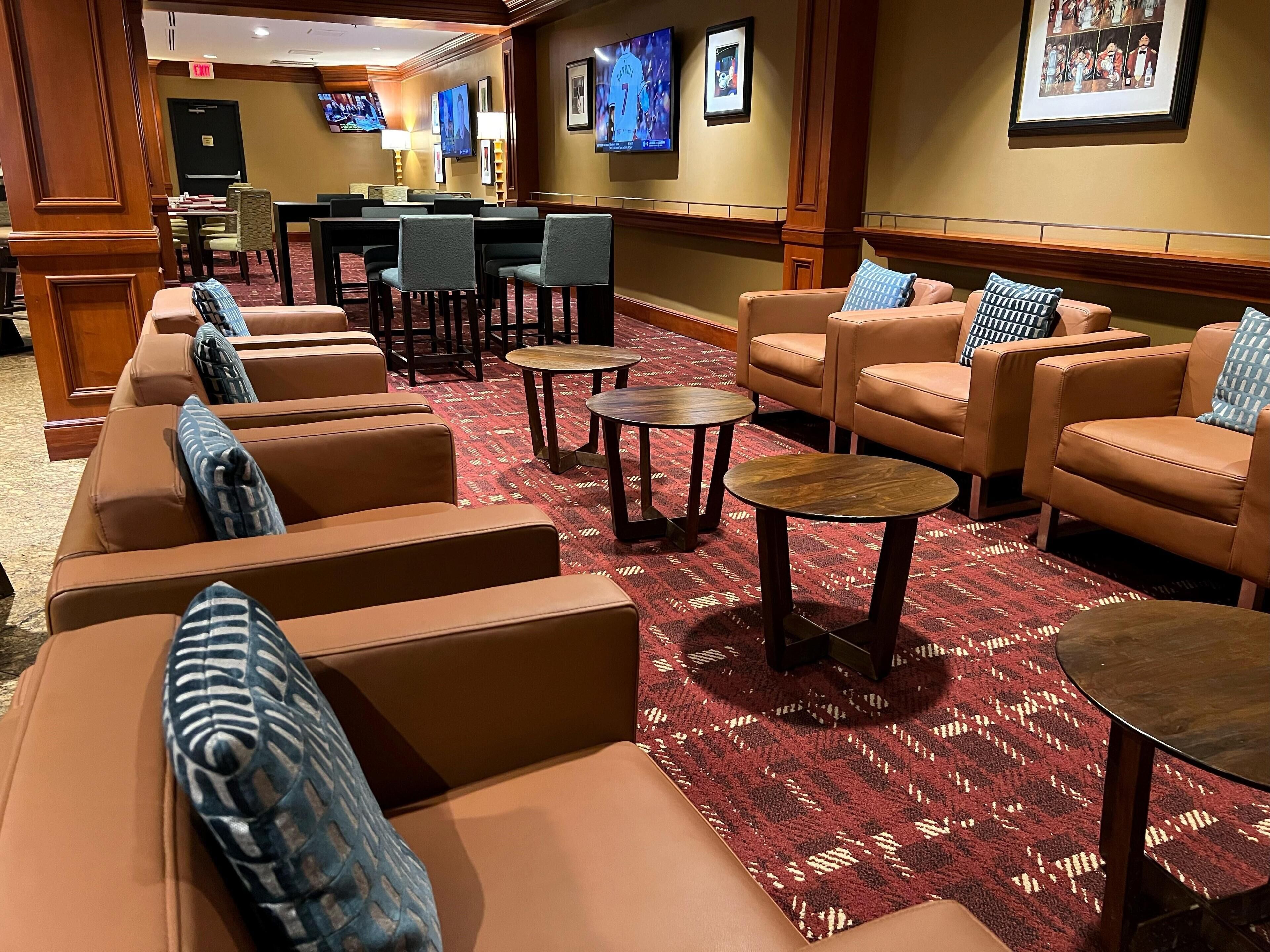 A hotel lobby with leather sofas, armchairs, and small round tables. There are framed pictures on the walls and a TV mounted on the wall.