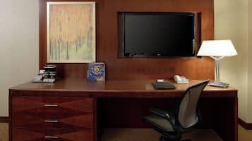 A hotel room with a wooden desk, a flat-screen TV, a lamp, a chair, and a painting on the wall.