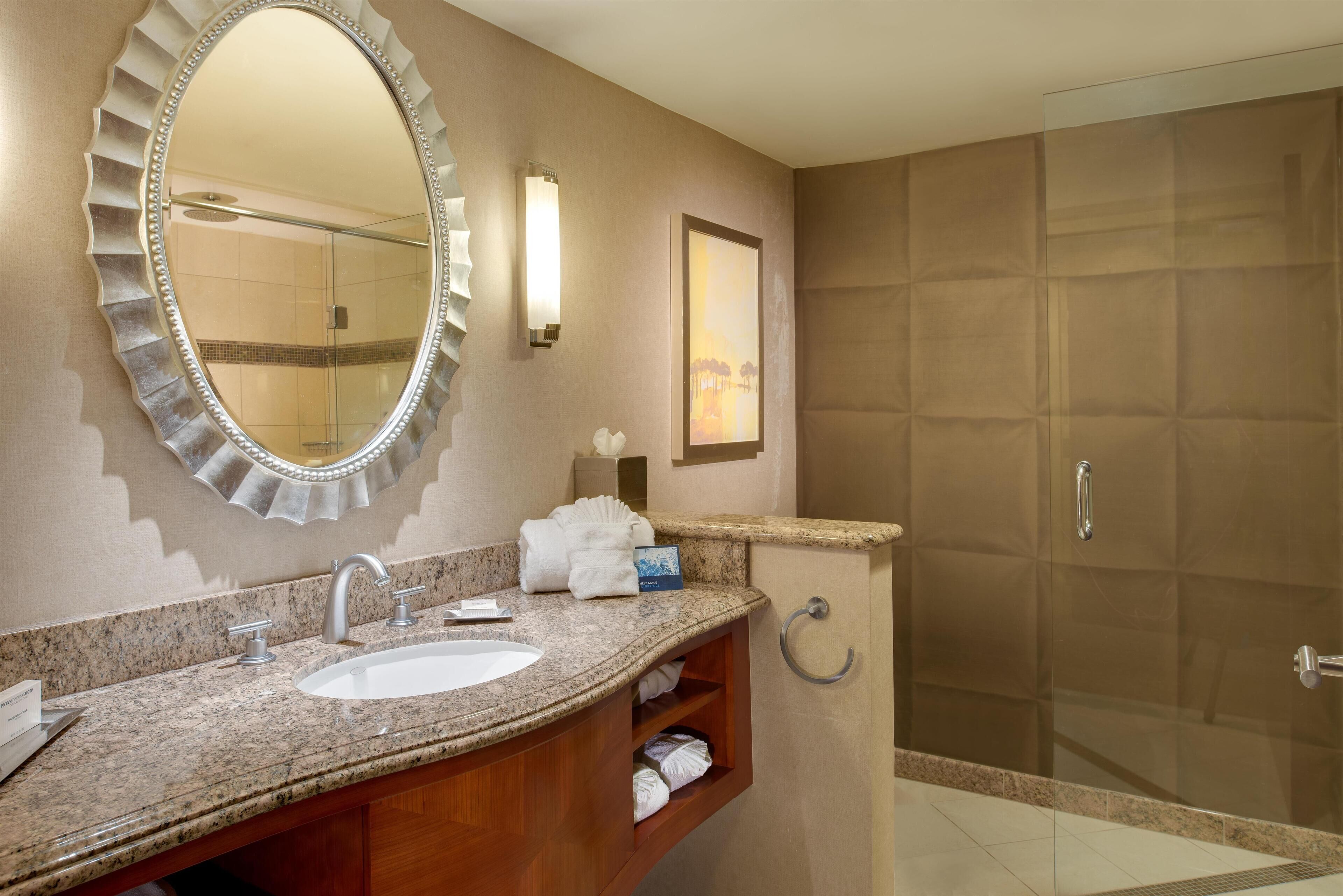 A bathroom with a large oval mirror, a granite countertop sink, a wooden vanity, a towel rack, and a glass-enclosed shower area.