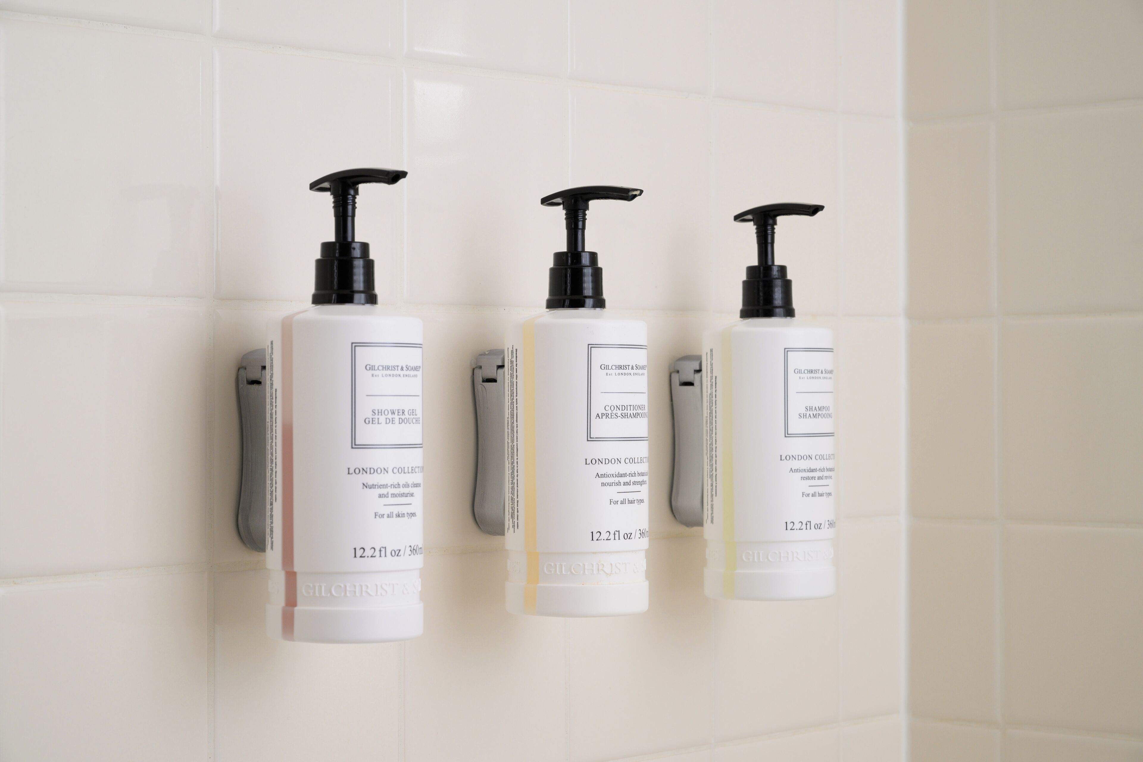 Trois flacons de shampoing de la marque Gilchrist & Soar sont fixés sur un mur recouvert de carreaux blancs.