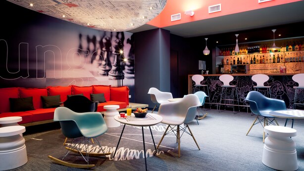 Een moderne zithoek met een rode bank, witte en blauwe stoelen, een klein tafeltje met een mand croissants, en een bar met diverse flessen.