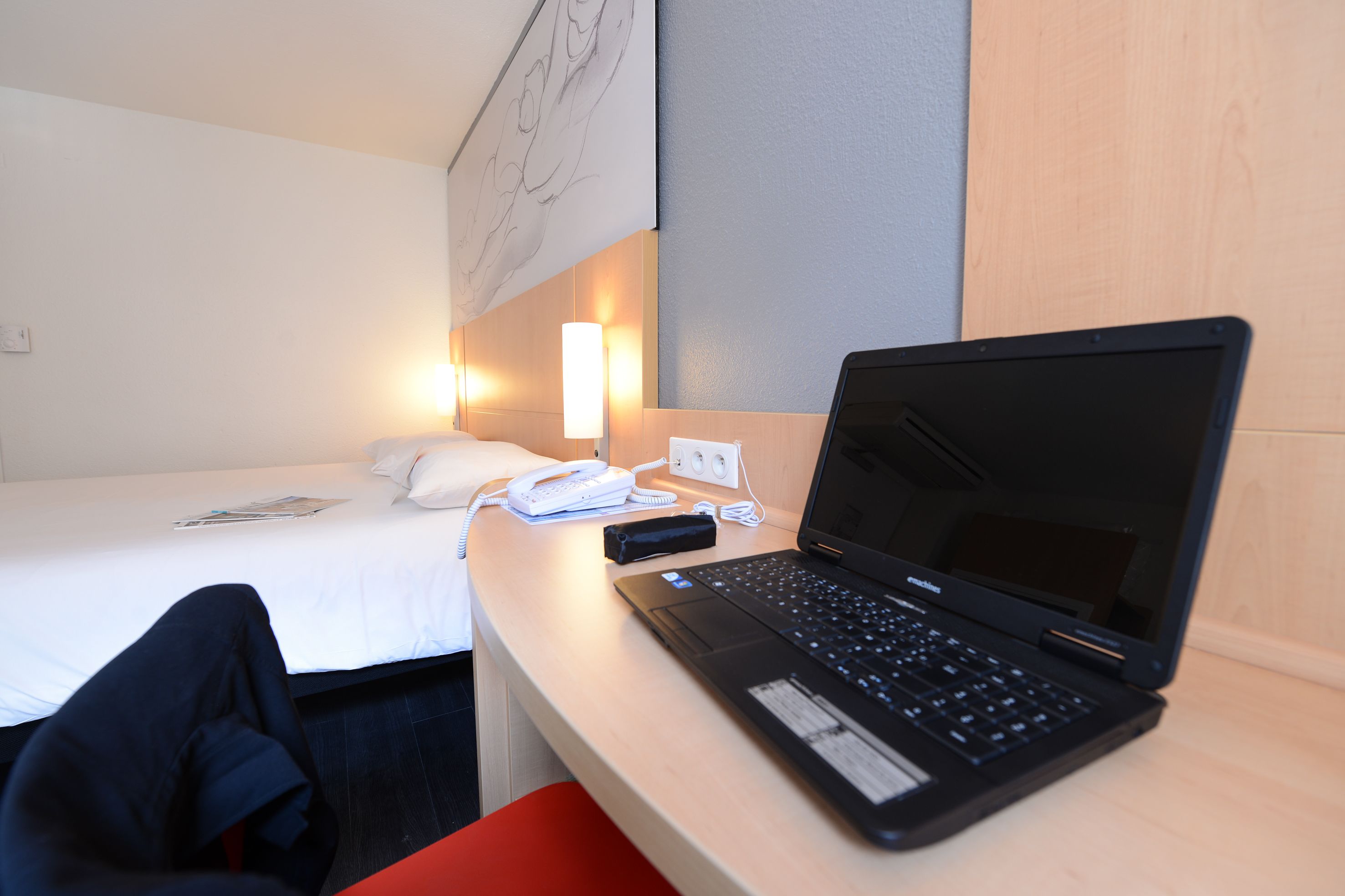 A hotel room with a bed, a desk with a laptop, a phone, and a wall-mounted light.
