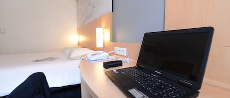 A hotel room with a bed, a desk with a laptop, a phone, and a wall-mounted light.