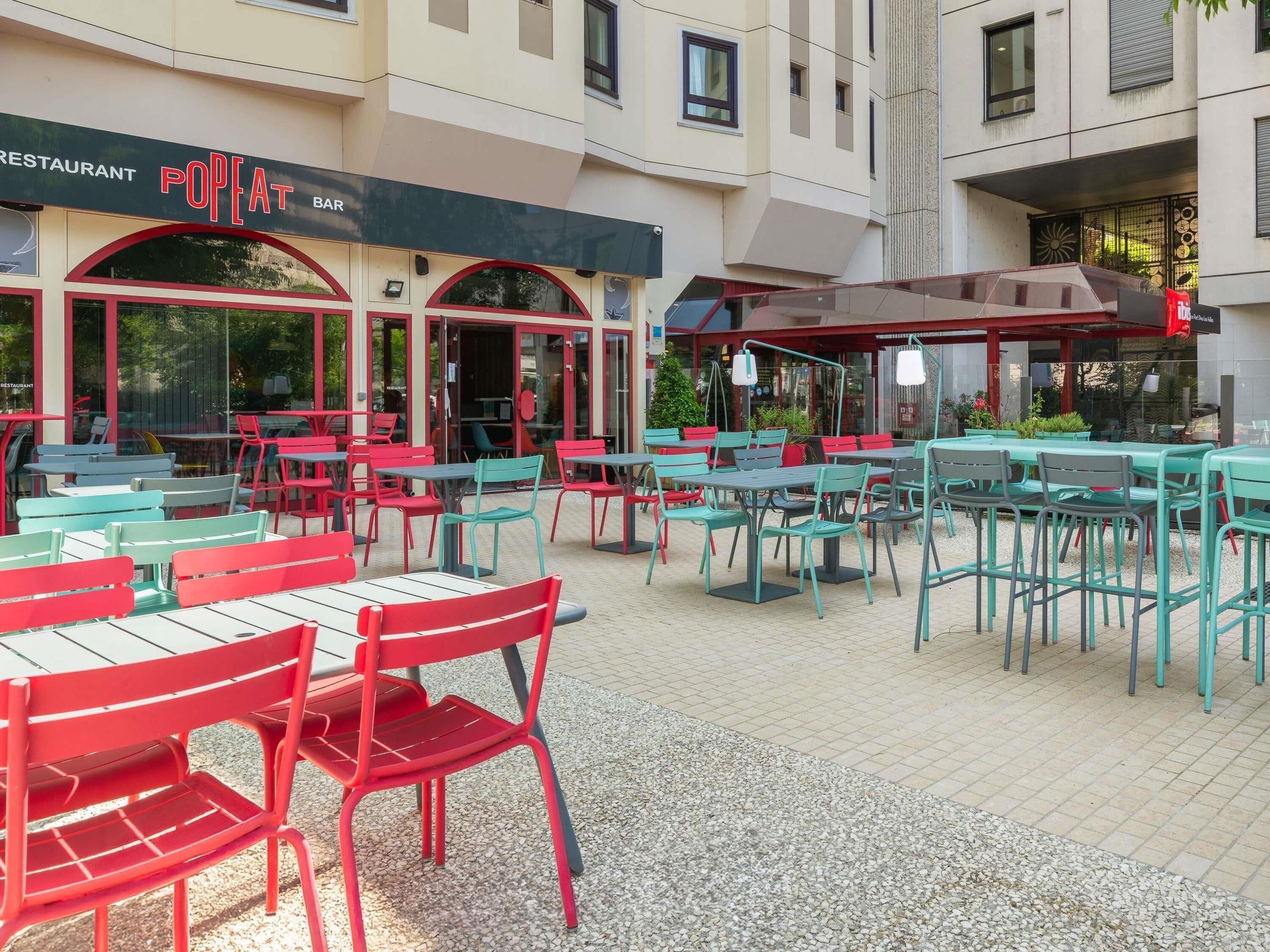 Un área de estar al aire libre con mesas y sillas, un restaurante llamado 'POPEAT BAR' al fondo, y la fachada de un edificio.