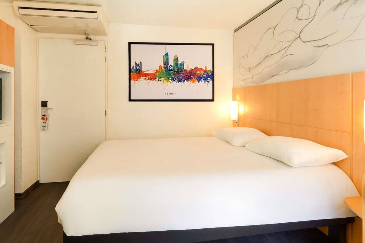 A hotel room with a bed, a framed cityscape artwork, a wall-mounted light, and a door with a fire alarm.