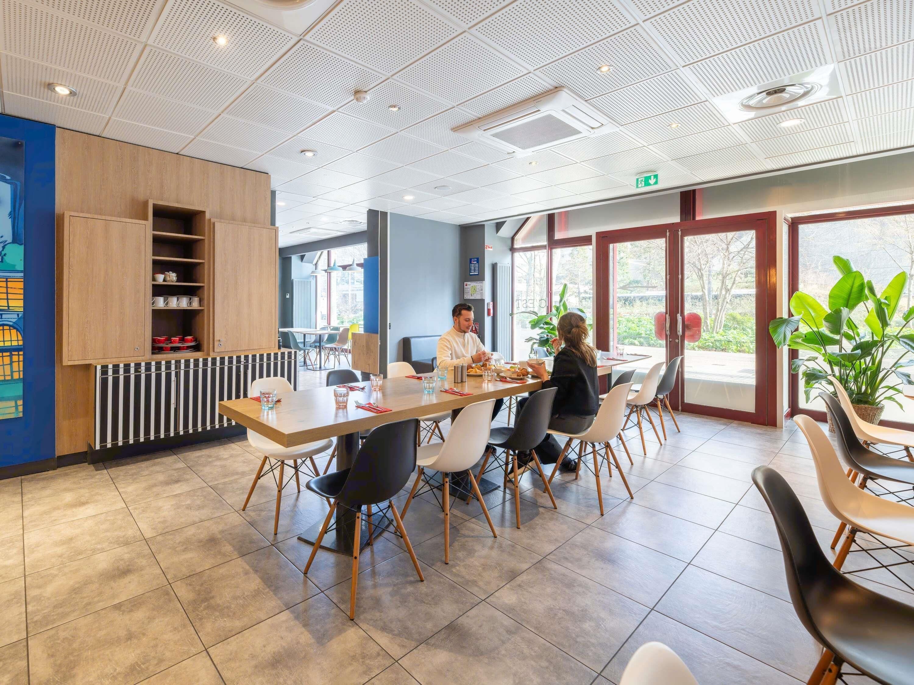 A modern dining area with a long table, chairs, and a view through large windows.