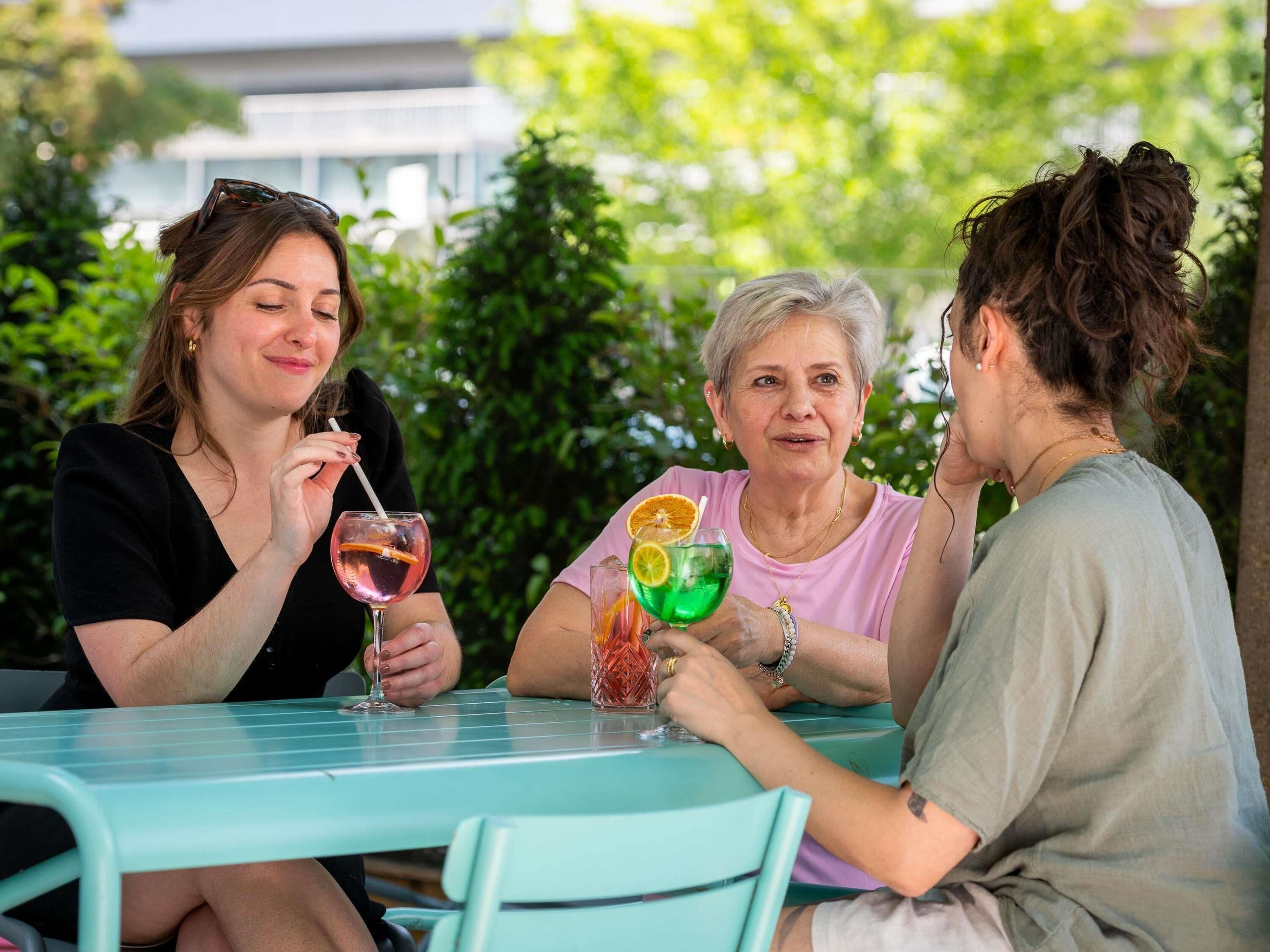 Tre persone sedute a un tavolo con bevande, all'aperto con il verde sullo sfondo.